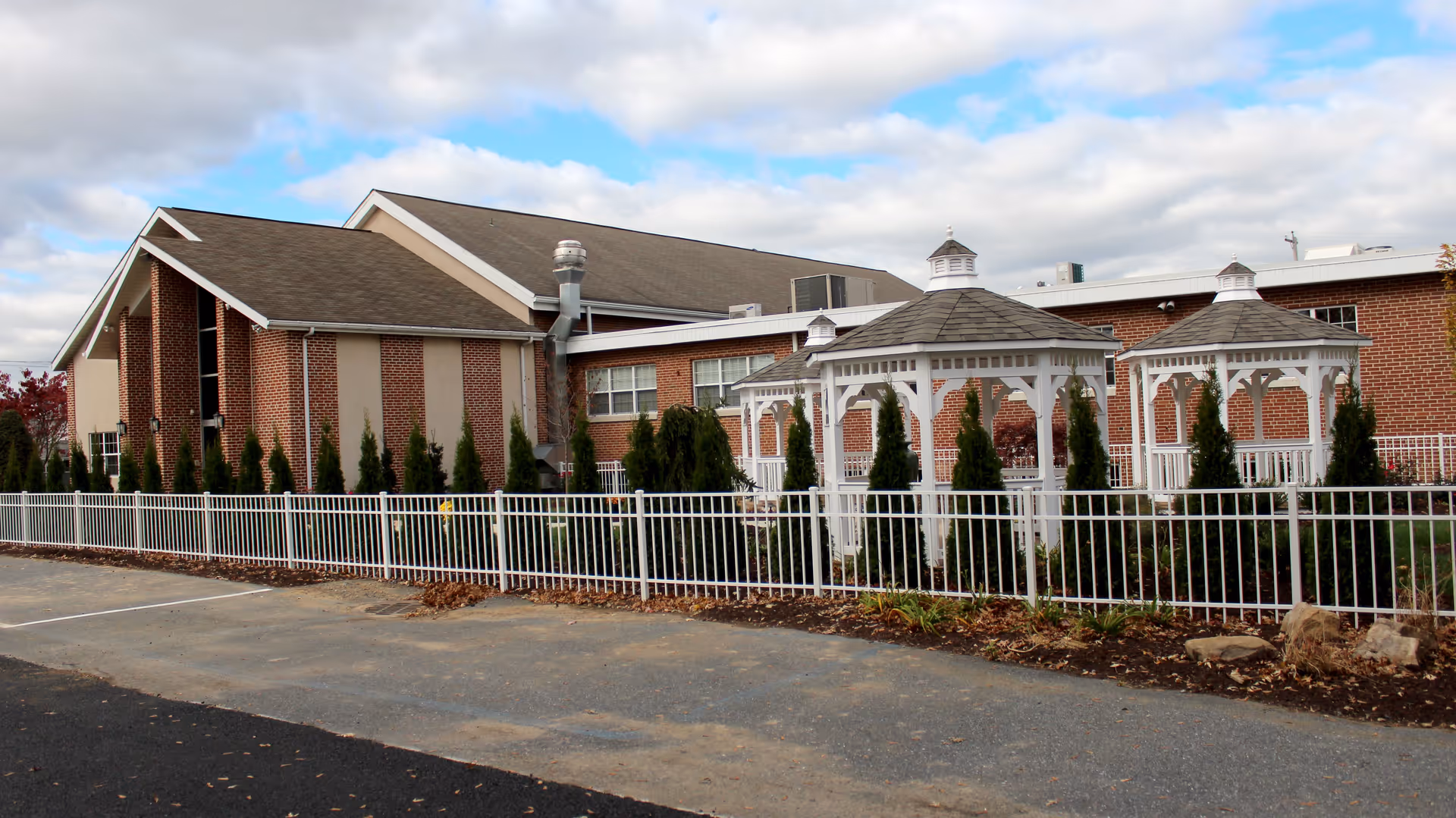 Exterior view of a brick building with a sloped roof, surrounded by a white metal fence. In front of the building are two white gazebos and several small evergreen trees. The sky is partly cloudy with patches of blue.