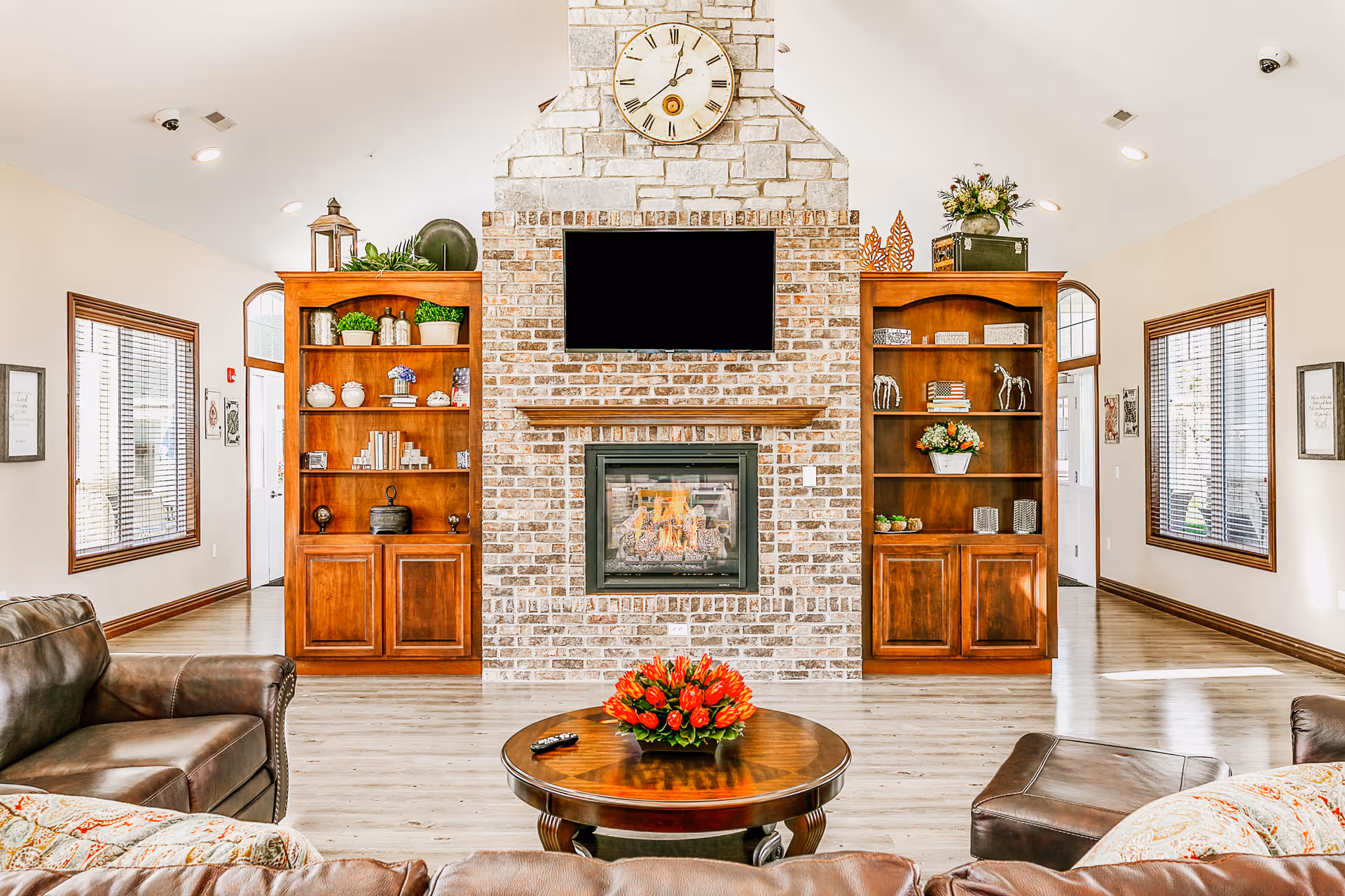 A cozy living room with a central brick fireplace and a mounted flat-screen TV above it. On either side of the fireplace are wooden built-in shelves decorated with plants, books, and decorative items. A round wooden coffee table with a red flower arrangement and a TV remote is in front of brown leather sofas and an ottoman. The room has large windows with wooden blinds and light-colored walls and flooring.