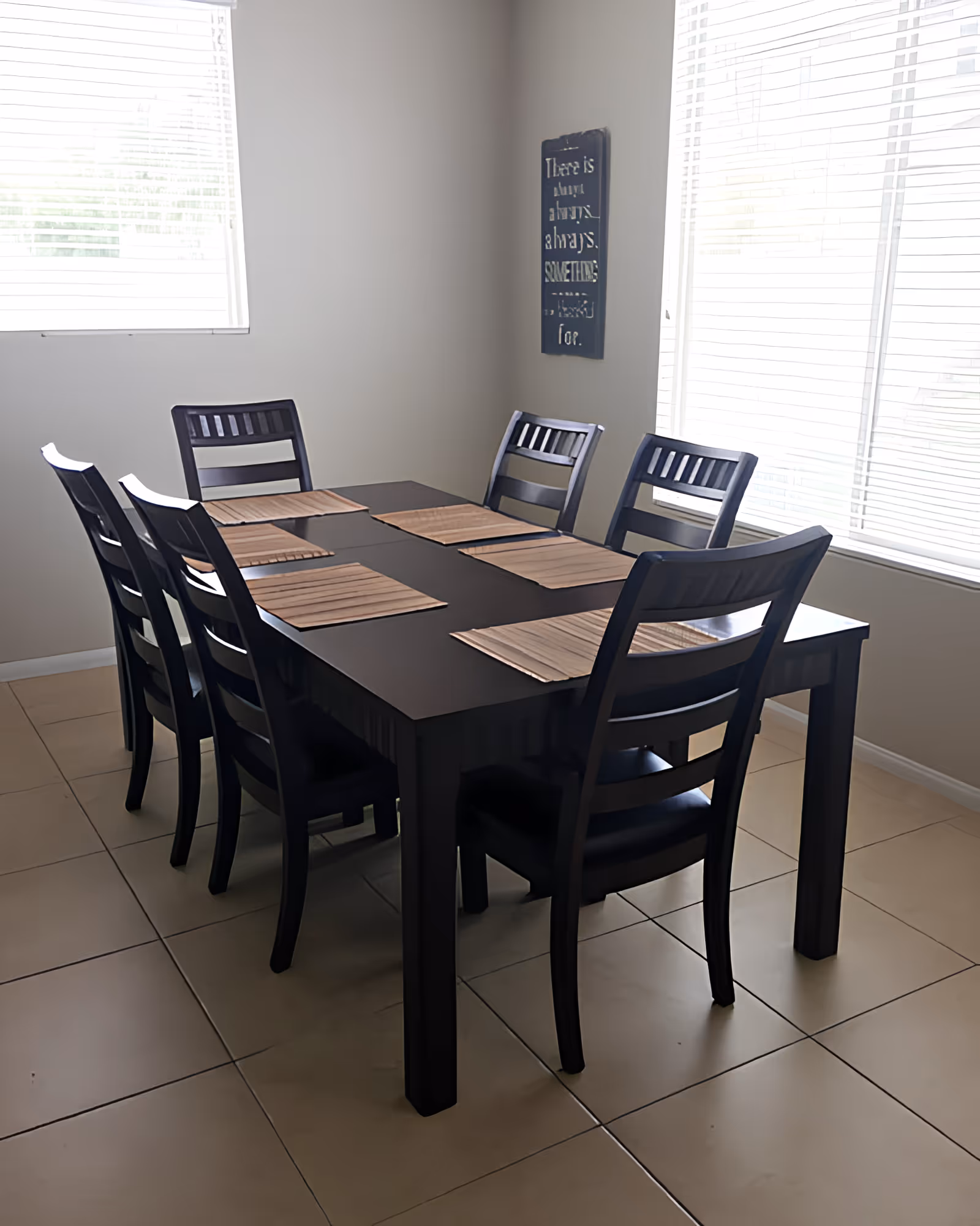 Bright dining room with a rectangular dark wood table, six matching chairs, and placemats by two windows.