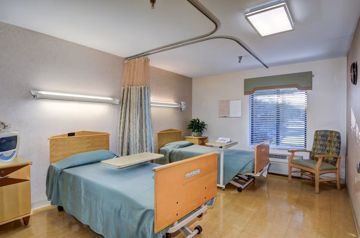 A hospital-style patient room with two adjustable beds, curtains, and a windowed seating area.