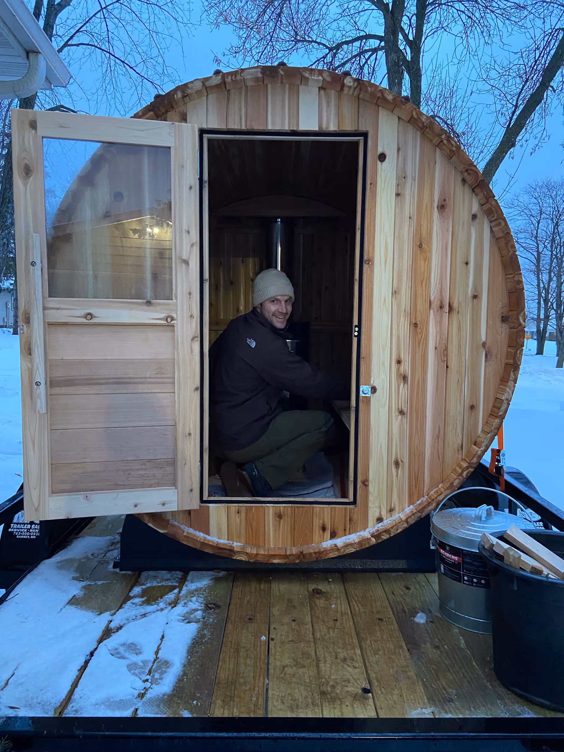 A wooden barrel sauna on a trailer outdoors, door open, a man sits inside.