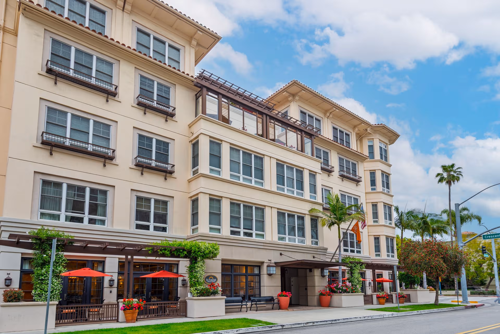 Multi-story beige senior living building with storefronts, red umbrellas, and palms along a sunny street.