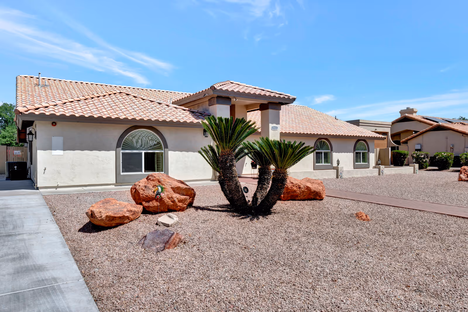 Single-story stucco building with a tiled roof and desert landscaping with large rocks and sago palm plants in the front yard.