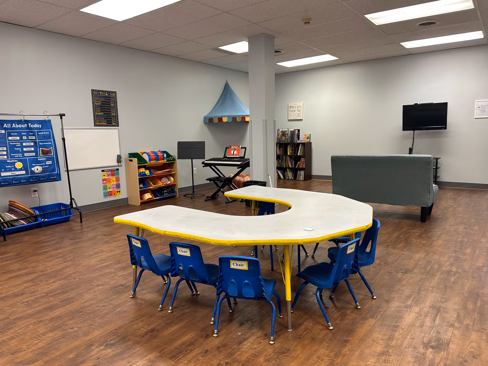 Spacious indoor activity room with a curved table, blue chairs, shelves, and a TV.