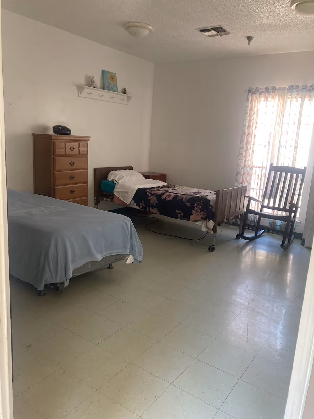 Simple shared bedroom with two single beds (one on wheels), a wooden dresser, and a rocking chair by a curtained window.