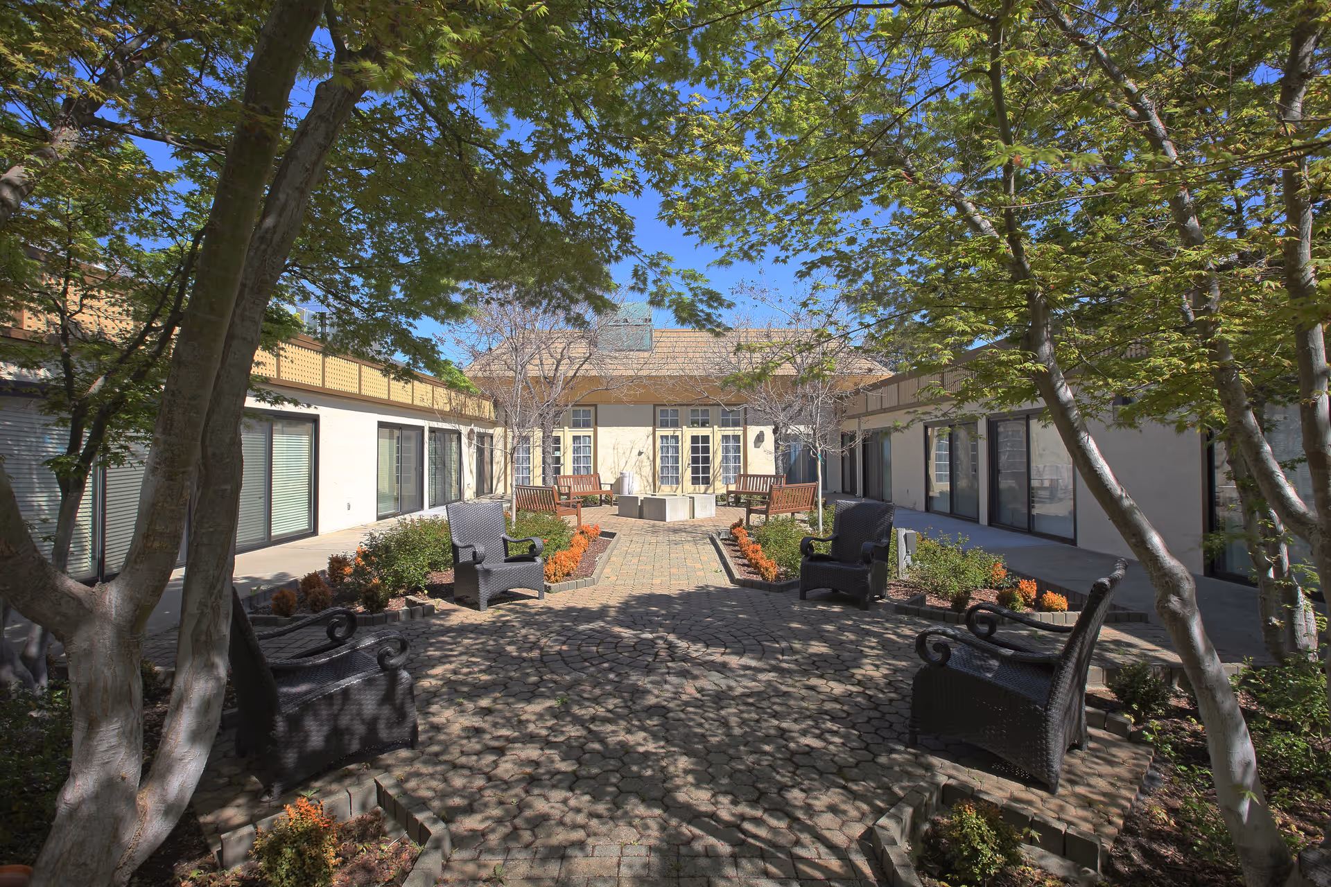 Outdoor courtyard area at Walnut Creek Skilled Nursing & Rehabilitation Center with trees providing shade, several black wicker chairs arranged around a paved circular area, and benches near a central fire pit. The courtyard is surrounded by a single-story building with large windows and doors.