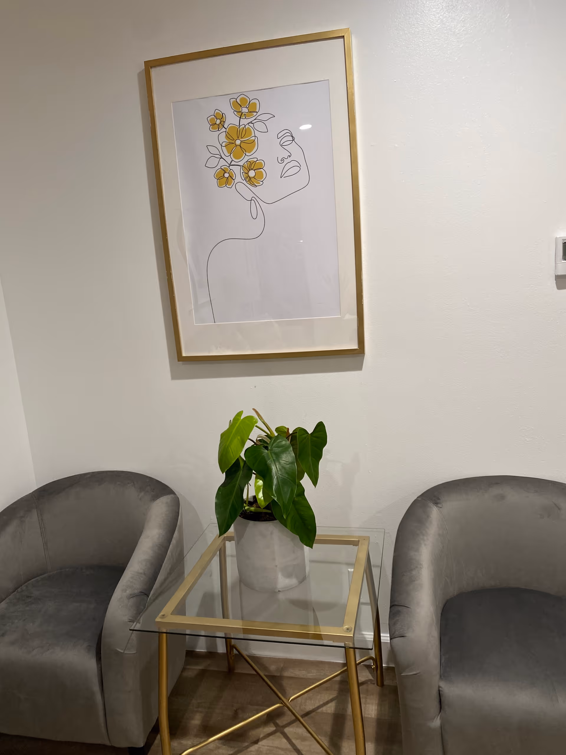Two gray chairs flank a glass-topped gold-framed side table with a potted plant.