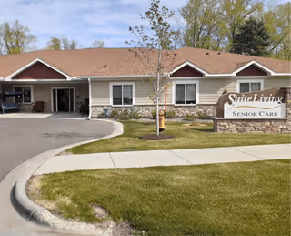 Front exterior of Suite Living senior care building with an entrance canopy, landscaped lawn, and a sign reading 'Suite Living Senior Care'.