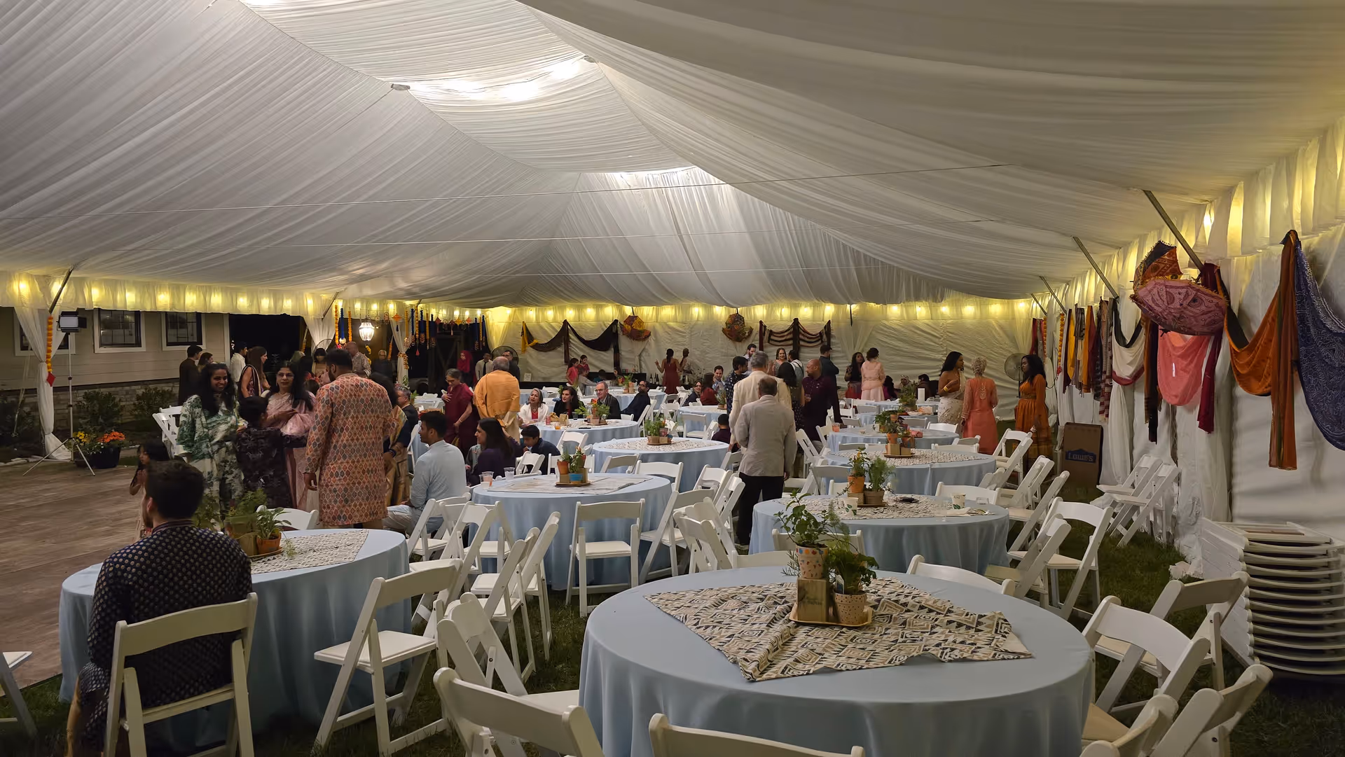 Outdoor tented reception with round tables, chairs, decor, and guests mingling.