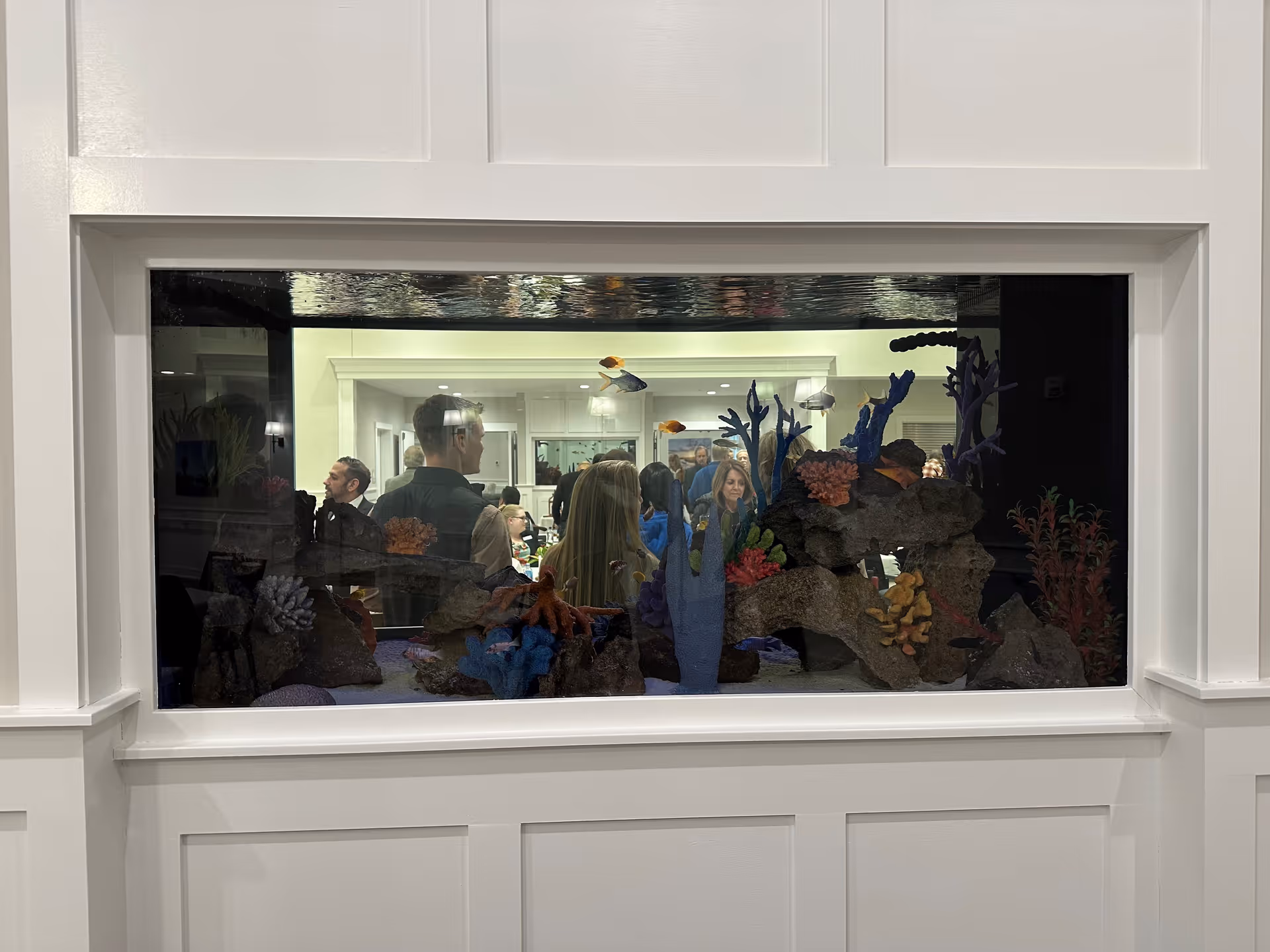 A large built-in aquarium with colorful coral and fish is set into a white paneled wall. Behind the aquarium, a group of people are gathered in a well-lit room, visible through the glass.