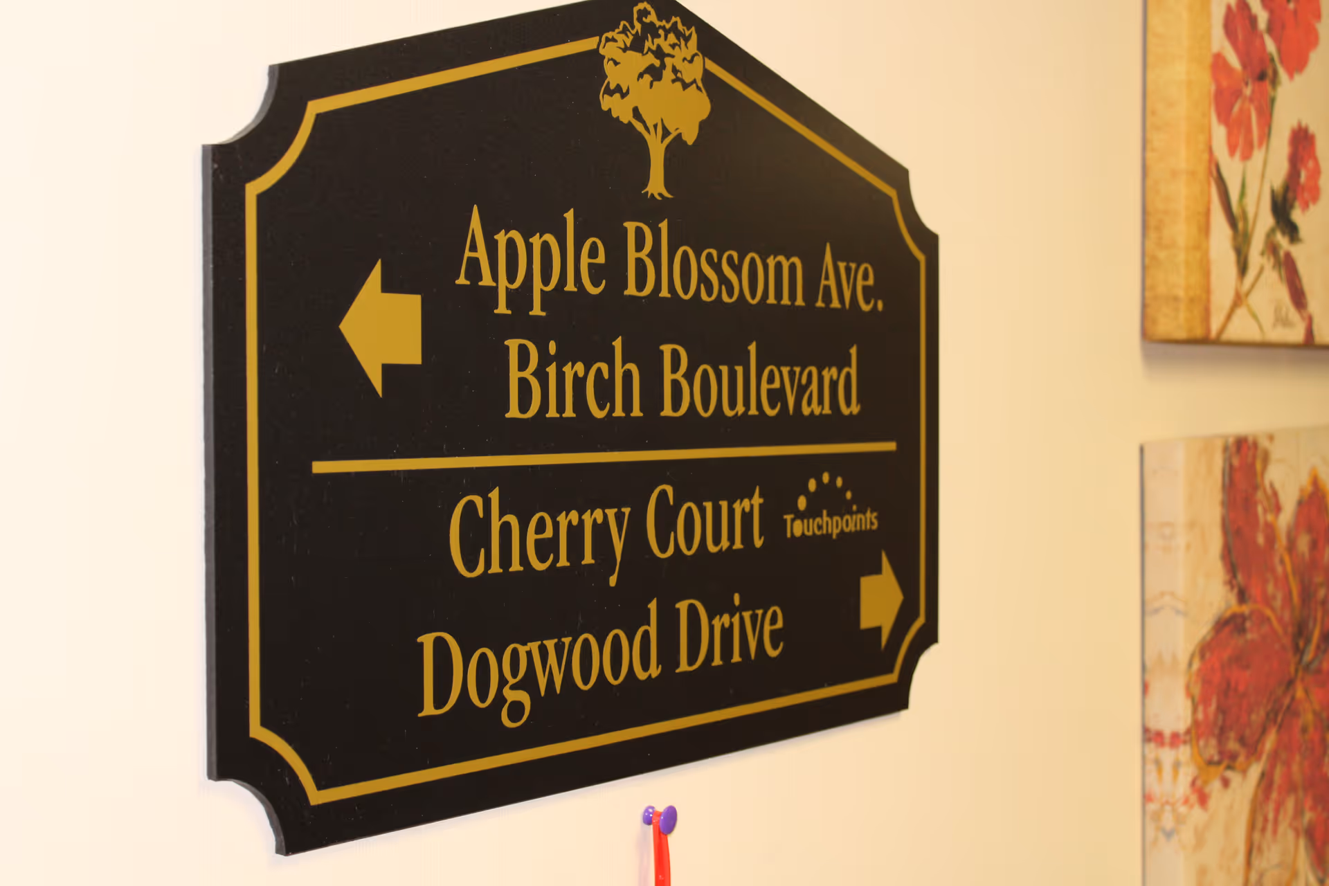 A black and gold directional sign mounted on a wall with arrows pointing left and right. The sign lists Apple Blossom Ave. and Birch Boulevard with a left arrow, and Cherry Court and Dogwood Drive with a right arrow. The word 'Touchpoints' and a small logo are also visible on the sign. Part of two floral paintings are visible on the right side of the image.