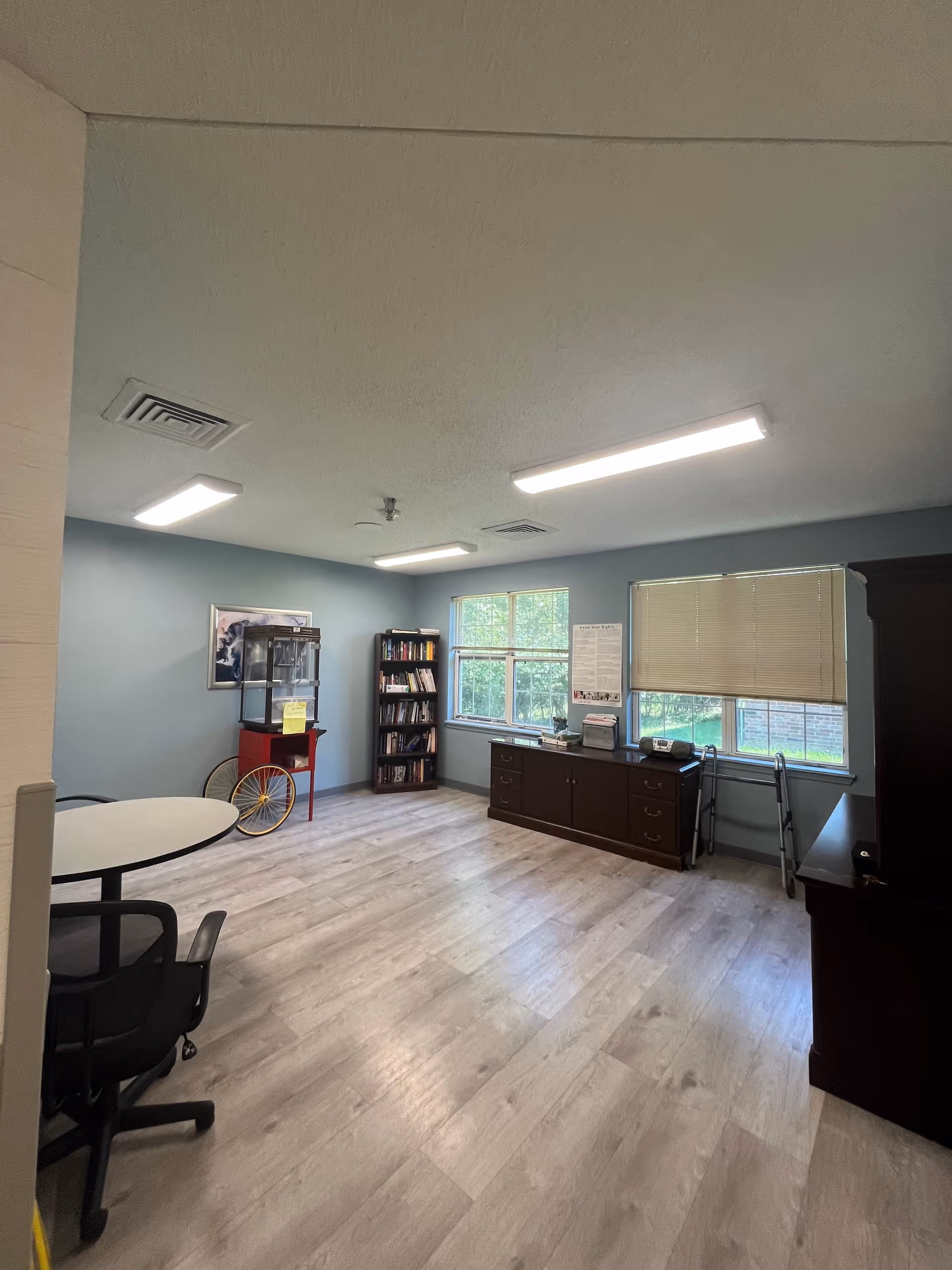 Interior community room with a round table, chairs, bookshelves, and a long cabinet.