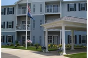 Multi-story senior living building with a covered entrance, balconies, flags, and landscaping.