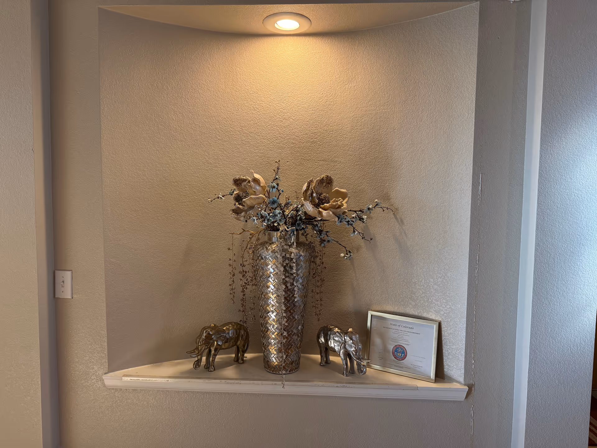 Decorative recessed alcove with a tall mosaic vase, dried flowers, elephant figurines, and plaque.