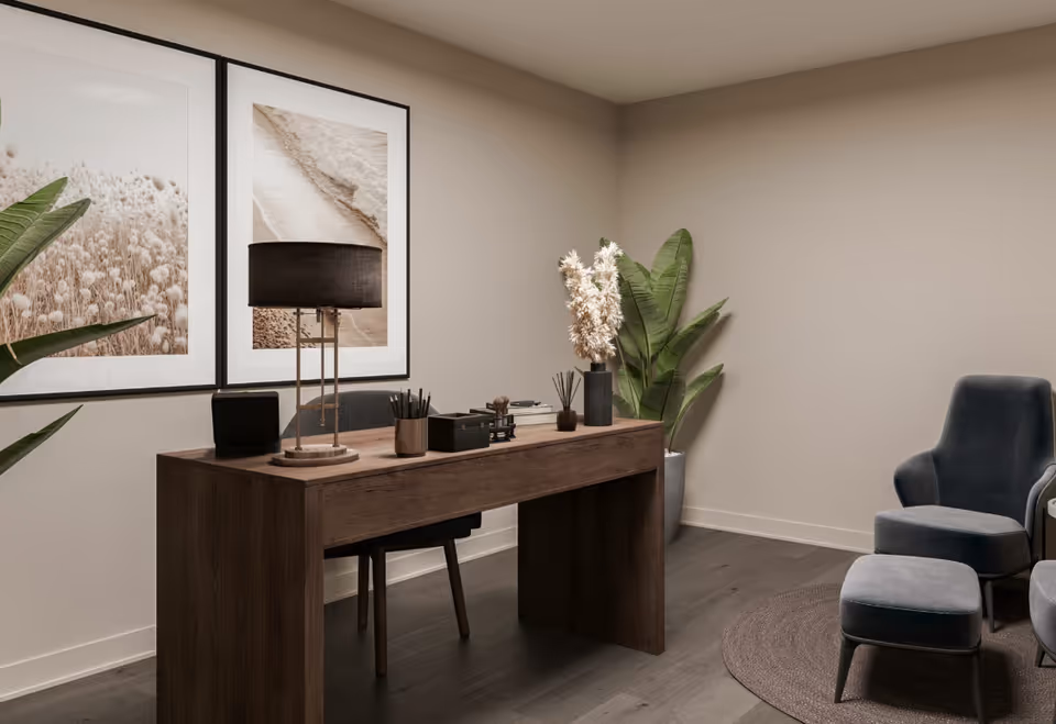 A modern office/sitting area with a wooden desk, table lamp, framed artwork, potted plants and upholstered chairs.