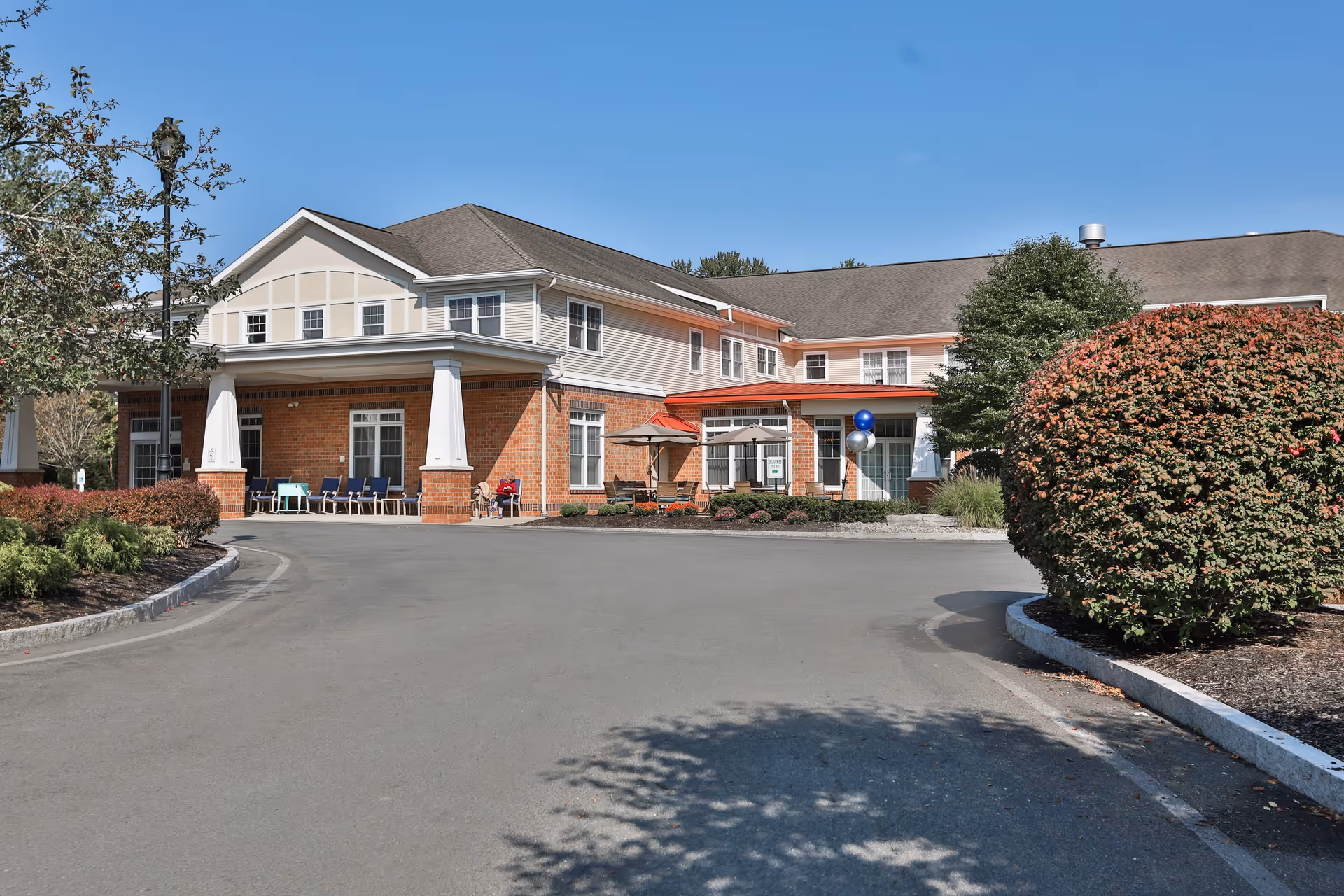 Exterior view of Peregrine Senior Living at Delmar Place, a two-story assisted living facility with a brick and beige siding facade, a covered entrance with white columns, outdoor seating, and landscaped bushes and trees under a clear blue sky.