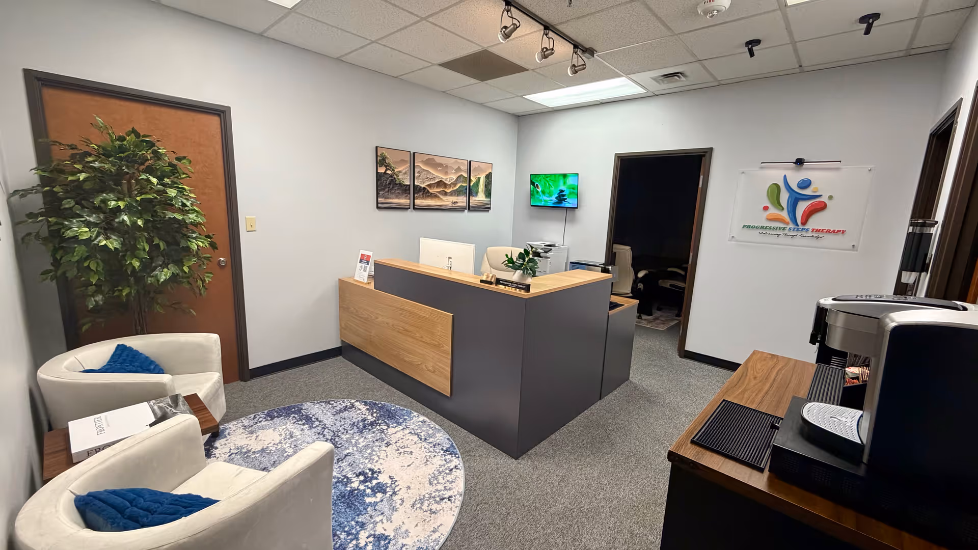 Reception area with a curved desk, seating, plants, wall artwork, and Progressive Steps Therapy sign.