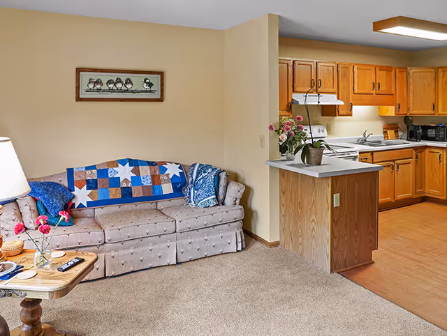 Open-concept living room with sofa, quilt, lamp, and adjoining wooden kitchen cabinets.