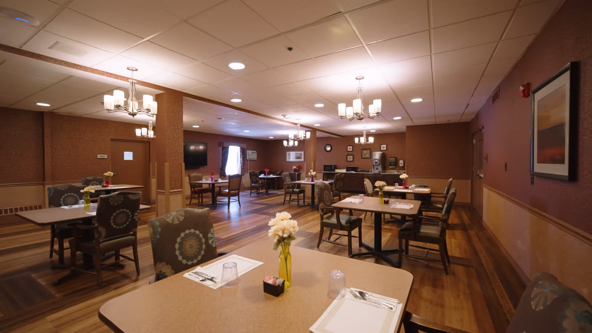 A spacious dining hall with round and square tables, chairs, and floral centerpieces.