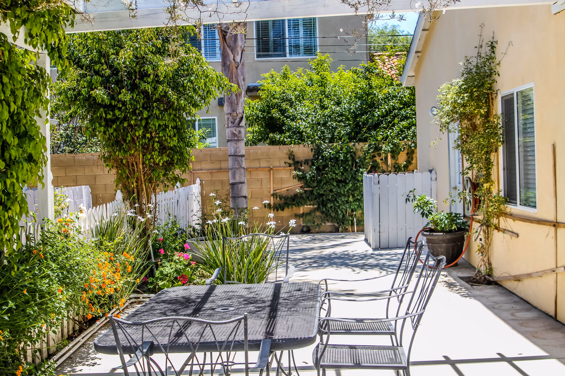 Sunlit outdoor patio with a metal table and chairs surrounded by potted plants, a white picket fence, and a beige building.