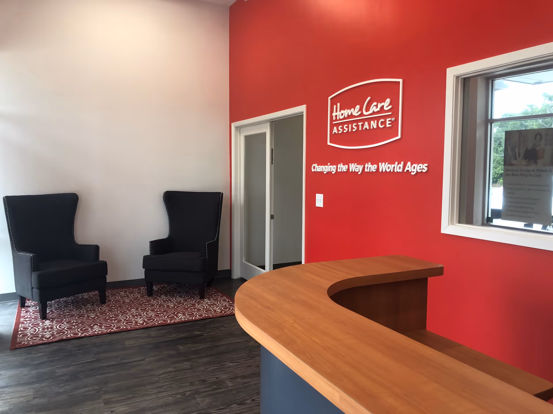 Reception area with curved wood desk, two black chairs, red wall with Home Care Assistance logo.