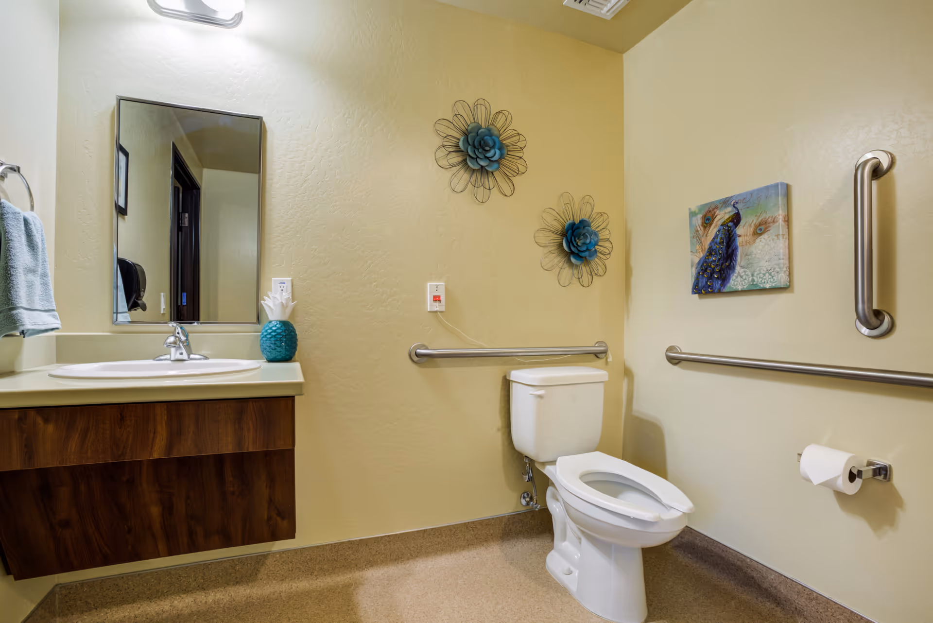 Bathroom with toilet, sink, mirror, grab bars, and wall art in a memory care facility