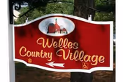 Outdoor red sign reading Welles Country Village with arrow, near trees.