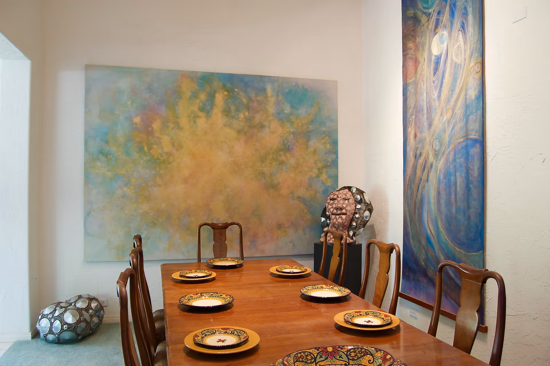 A dining room with a wooden table set with six decorative plates. Six wooden chairs surround the table. The walls feature two large abstract paintings, one with warm colors and the other with cool blue tones. There are two artistic sculptures placed on the floor and a pedestal near the paintings.