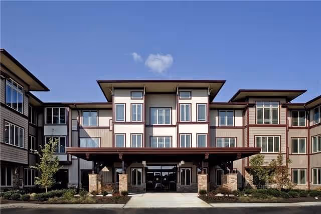 Three-story senior living building with a covered entrance and landscaped front under blue sky