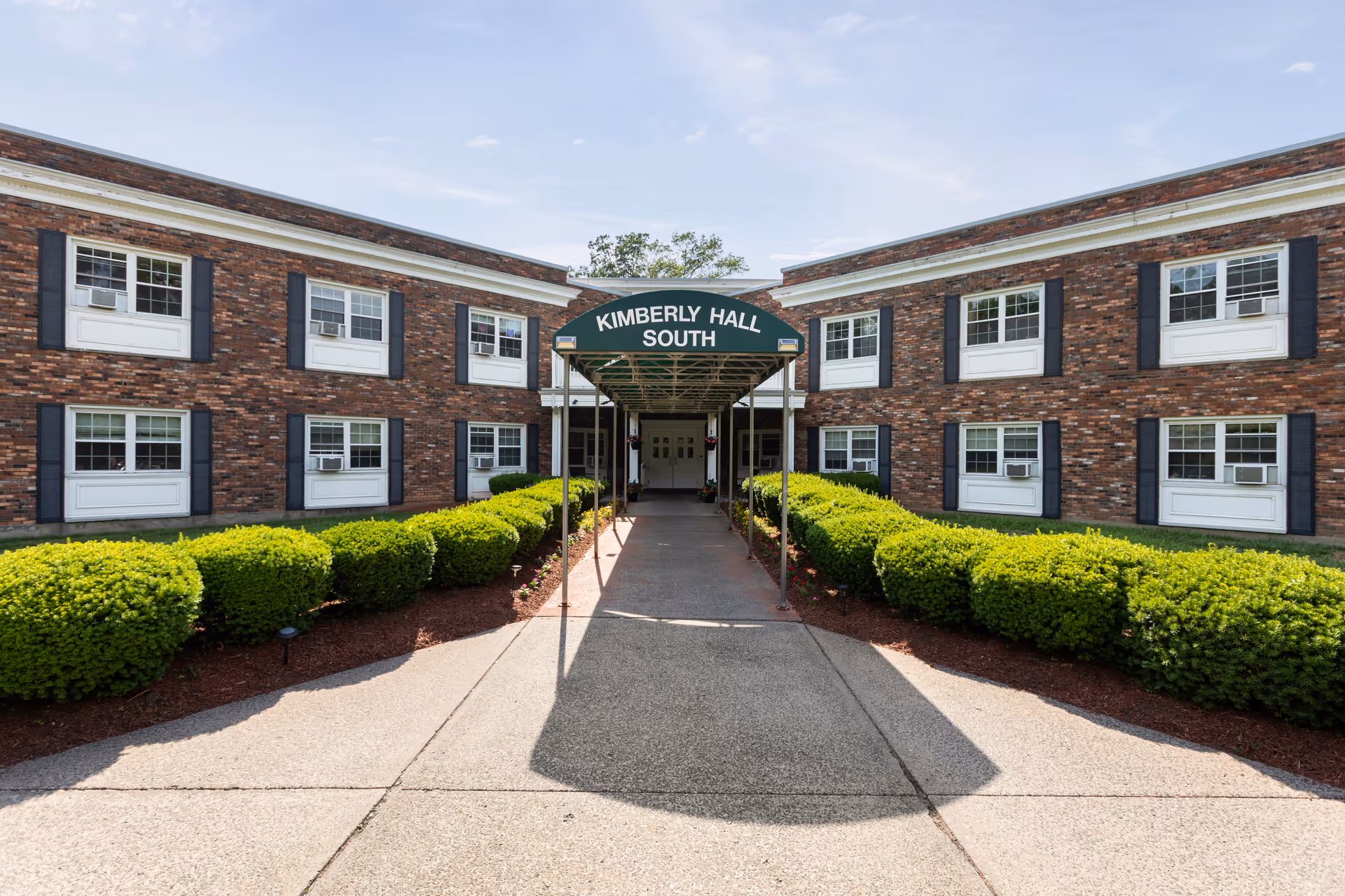 Exterior of Kimberly Hall South senior living building with a covered entrance and manicured shrubs.