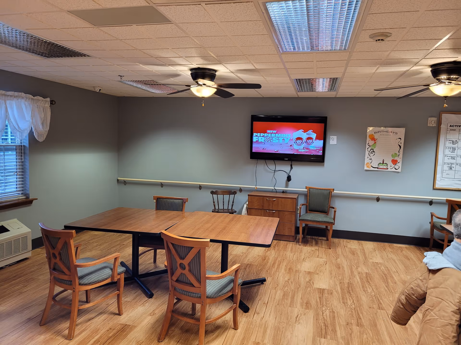 Senior living activity room with a TV, tables, and wooden chairs.