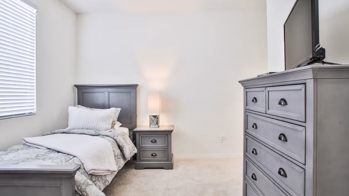 Minimalist bedroom with a single bed, nightstand with a lamp, and a tall dresser against a white wall.