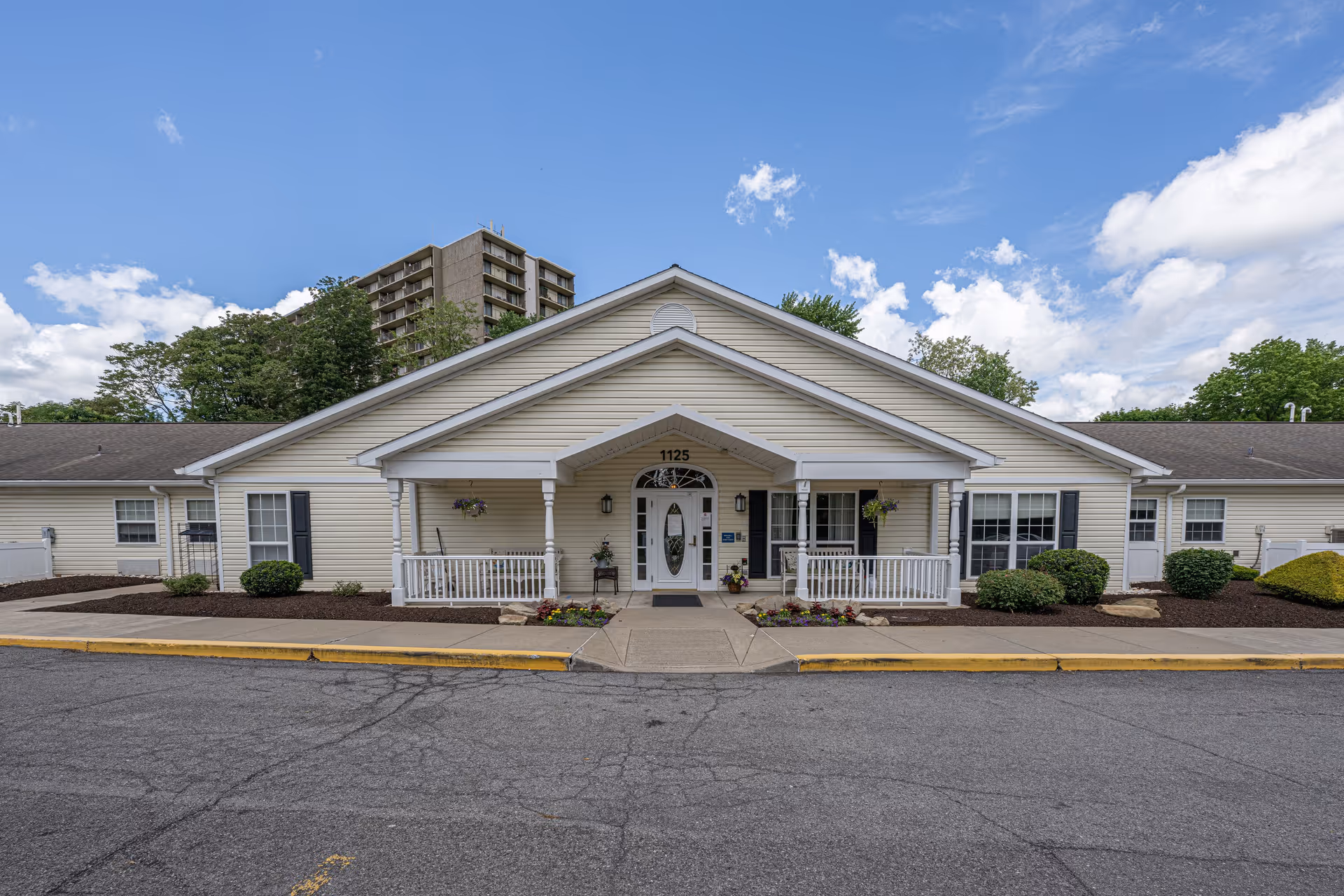 Front exterior view of a single-story building with beige siding, white trim, and a covered entrance with white railings. The building is surrounded by neatly maintained landscaping with bushes and flowers. The sky is partly cloudy with blue patches visible.