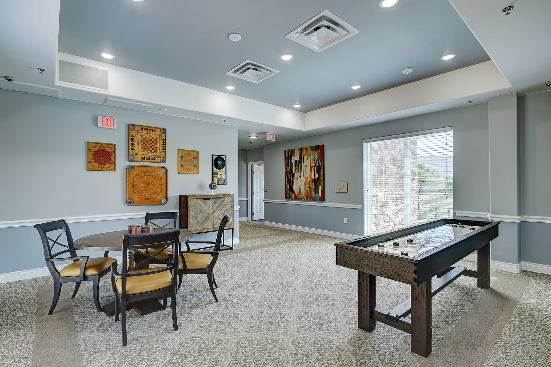 Bright senior living common area with round table, chairs, art, and a game table by windows.