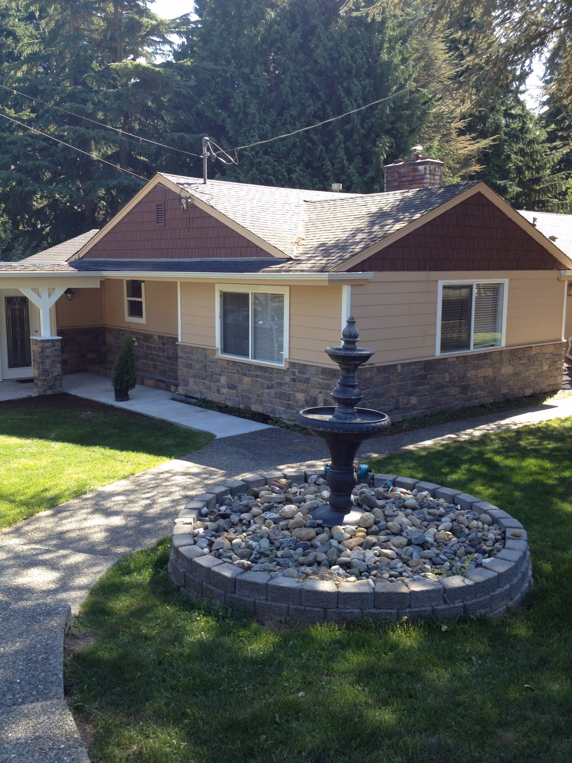 Exterior view of a single-story house with beige siding and stone accents. There is a small circular stone fountain filled with rocks in the front yard, surrounded by green grass and a concrete pathway leading to the entrance. Tall evergreen trees are visible in the background.