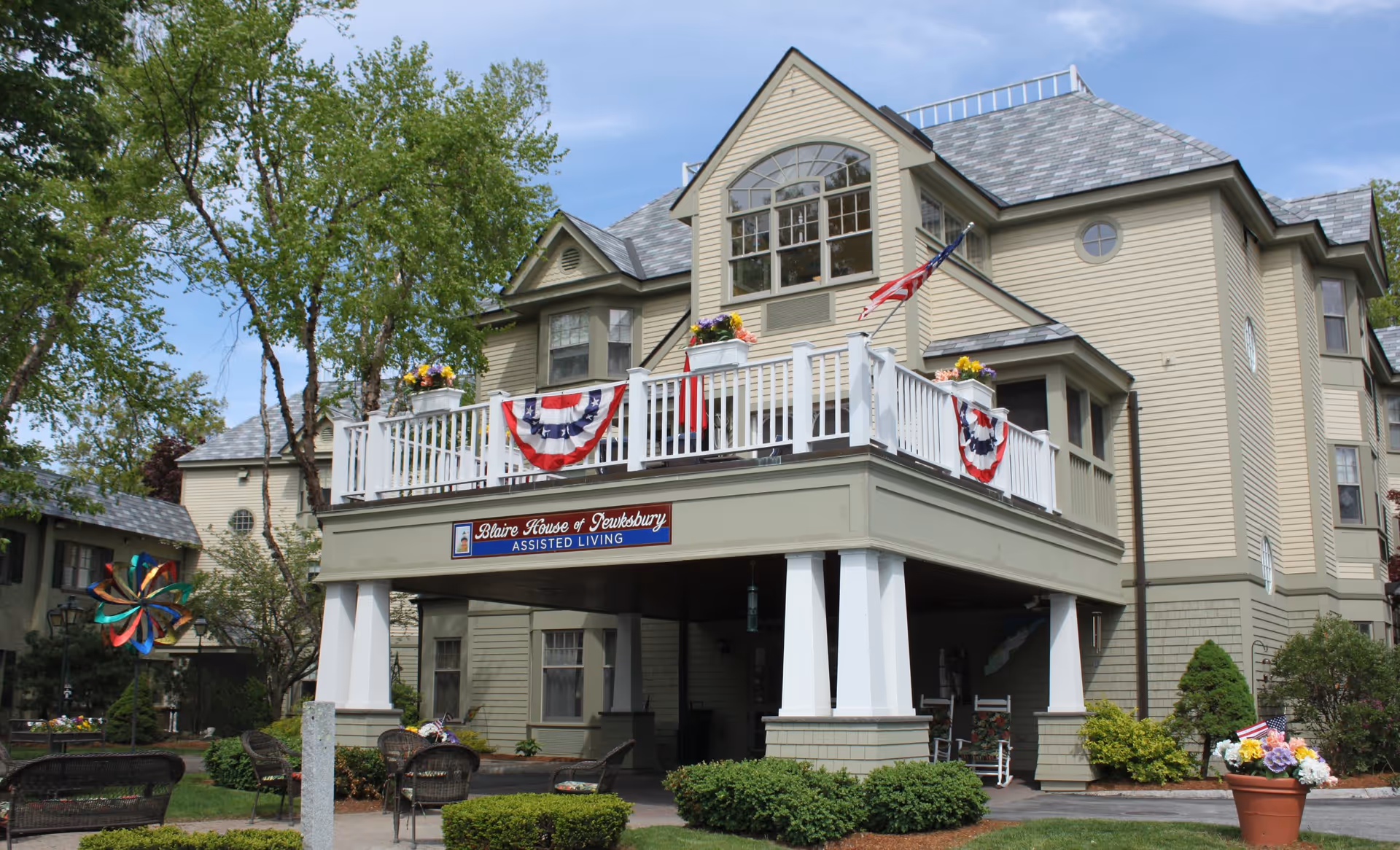 Exterior view of Blaire House of Tewksbury Assisted Living, a large beige multi-story building with a balcony decorated with patriotic bunting and flowers. There are trees, shrubs, and outdoor seating in the foreground under a clear sky.
