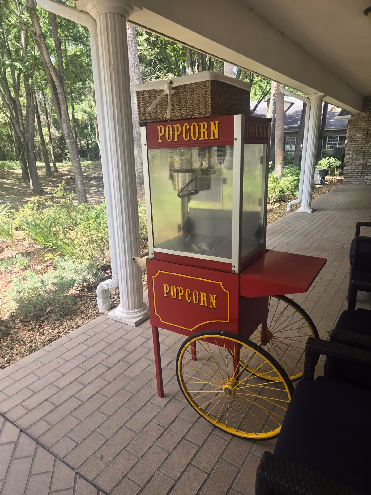 Red popcorn cart on a covered outdoor patio beside chairs and lush trees.