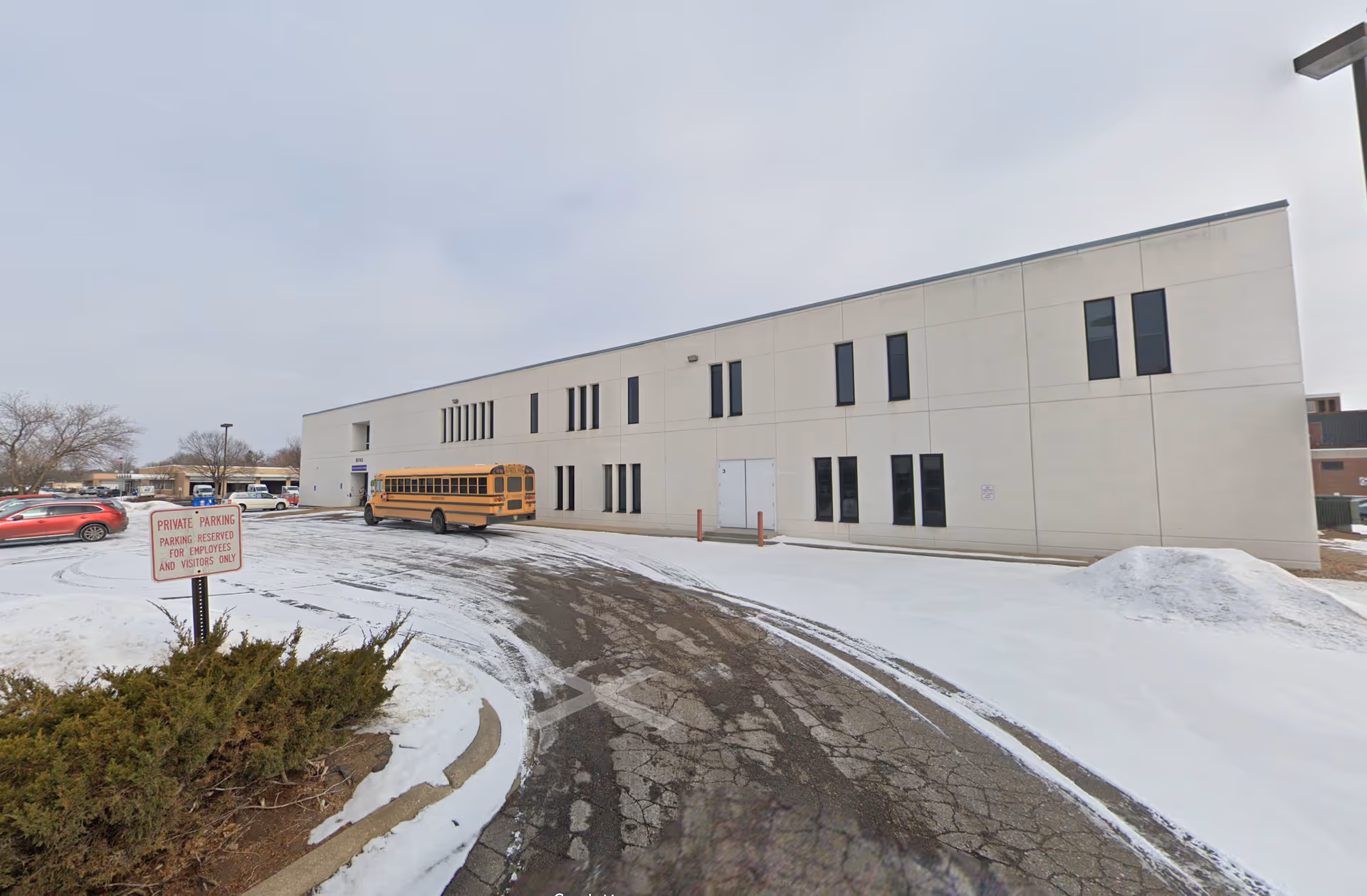 Exterior view of a beige facility with many windows, a yellow school bus, and snow-covered parking.