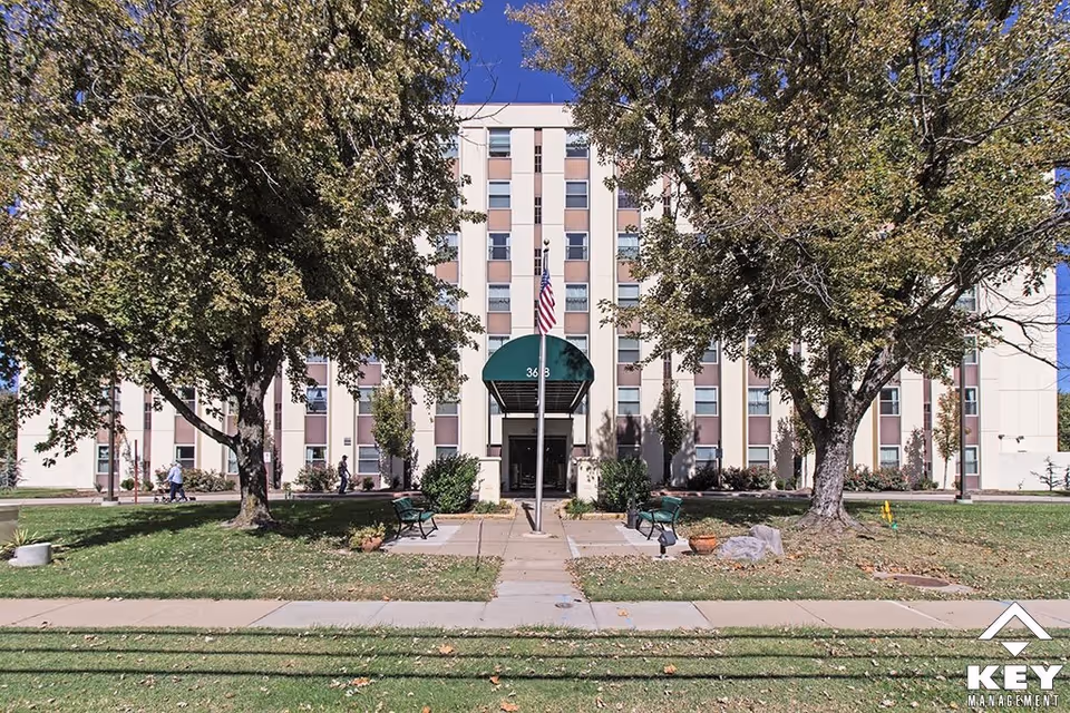 Front exterior of a multi-story senior living building with a green awning entrance, trees, benches, and an American flag.