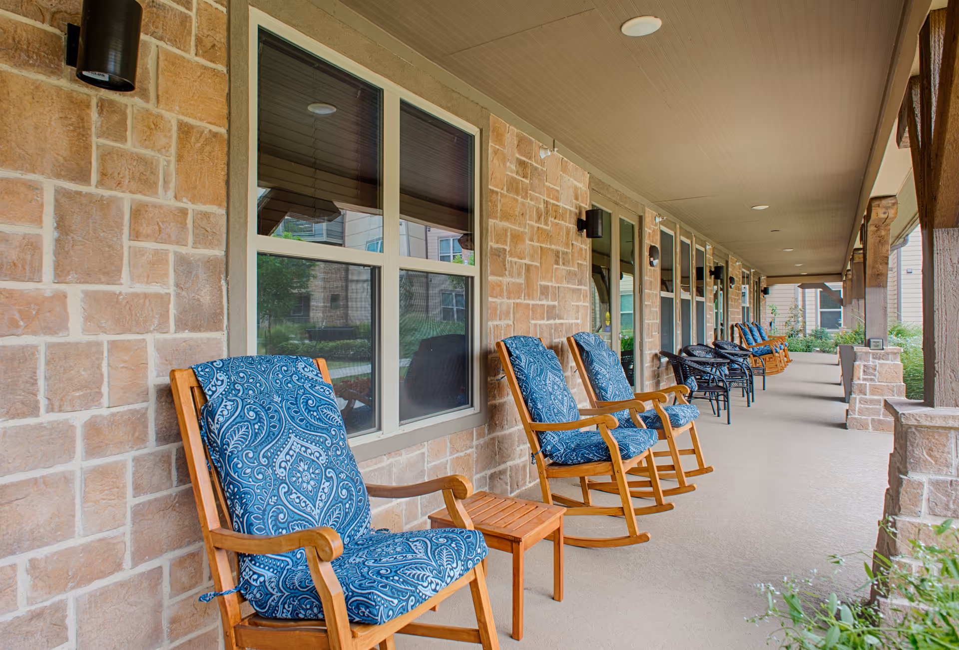 Row of cushioned rocking chairs along a covered outdoor walkway beside a brick building.