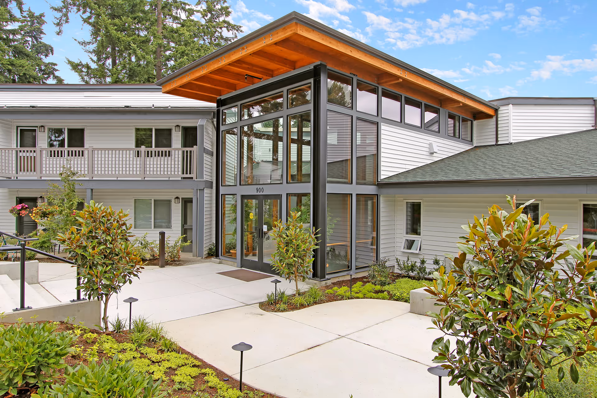Exterior view of Evergreen Court senior living facility showing a modern entrance with large glass windows, a wooden overhang roof, and surrounding greenery including bushes and small trees. The building has white siding and a second-floor balcony.