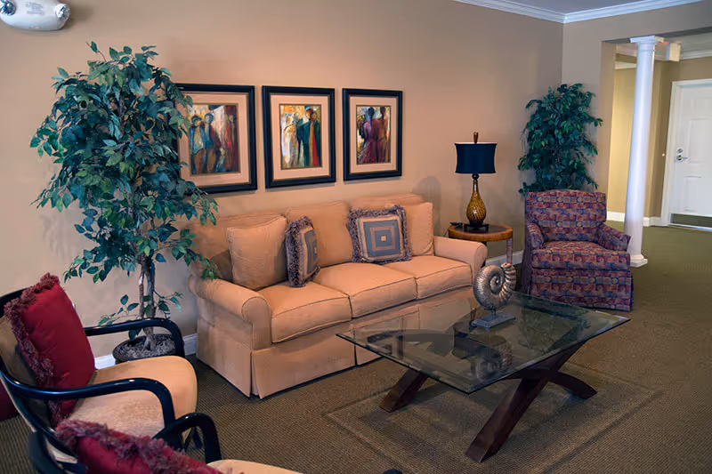 A cozy living room area in a retirement community featuring a beige sofa with decorative pillows, a glass coffee table with a decorative shell sculpture, two armchairs with red cushions, two large potted plants, a side table with a lamp, and three framed abstract paintings on the wall.
