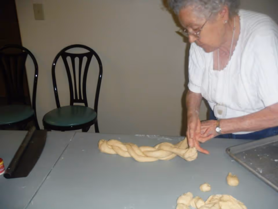 An elderly woman with gray hair and glasses is braiding dough on a table in a room with two black chairs with green seats in the background. There is a baking tray and some small pieces of dough on the table.