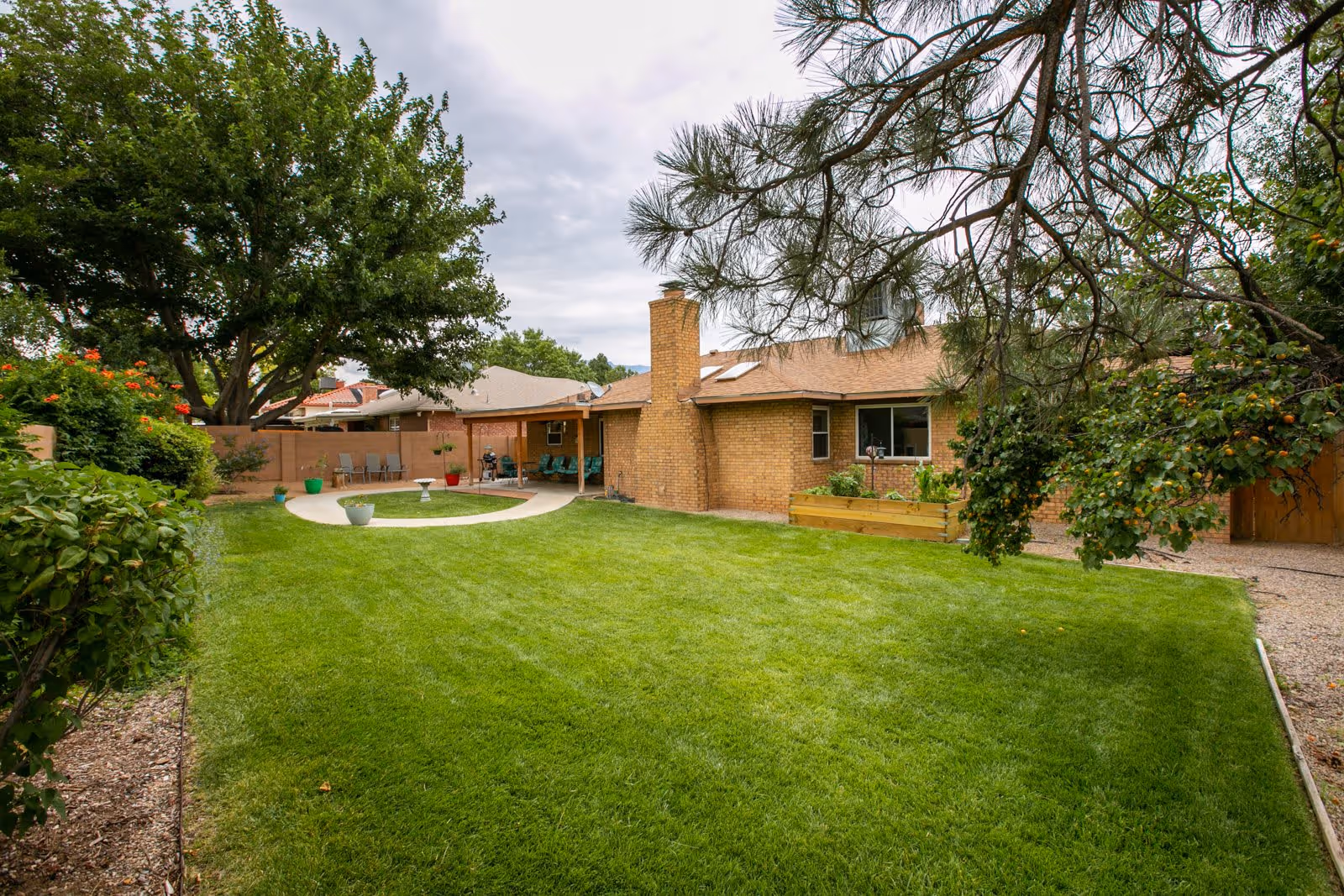 A spacious backyard with a well-maintained green lawn, surrounded by trees and shrubs. There is a circular concrete patio area with potted plants and outdoor seating near a brick building with a chimney. The sky is cloudy.