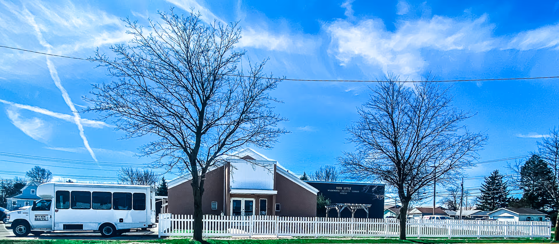 White bus parked beside a brown building with a white picket fence and leafless trees.