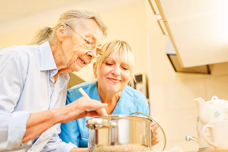 An elderly woman stirs a pot in a kitchen while a caregiver watches and smiles.
