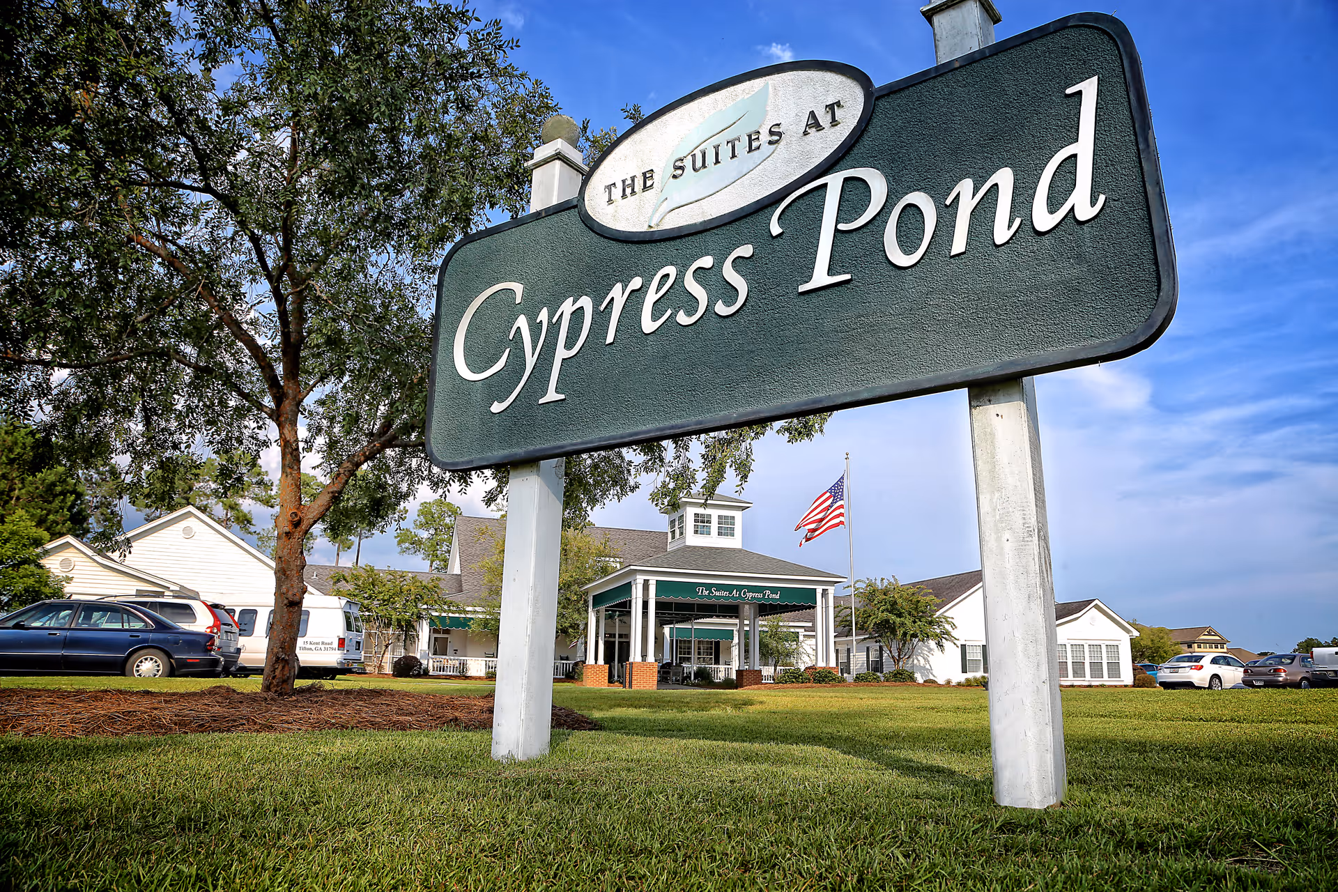 Large green sign reading 'The Suites at Cypress Pond' in front of a senior care facility building with an American flag, trees, parked cars, and a clear blue sky.
