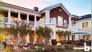 Exterior view of a senior living facility building at dusk with brick walls, white columns, balconies, outdoor seating areas with chairs and tables, umbrellas, and landscaped greenery.