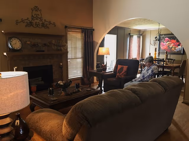 Living room in a senior care facility with a sofa, fireplace, and an elderly resident seated.