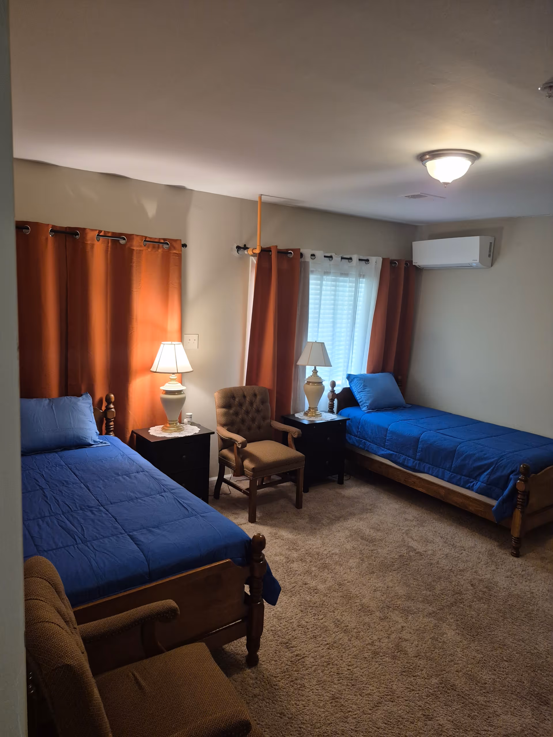 Two twin beds with blue comforters, lamps, nightstands, and an armchair in a carpeted bedroom.